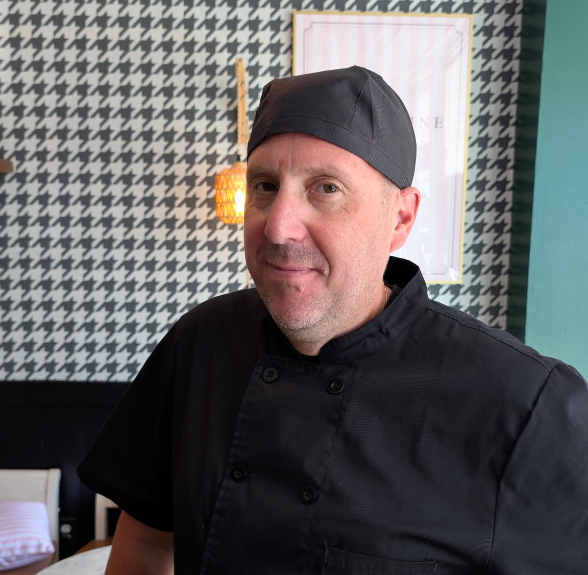 Arnaud, the chef at Madly Madeleine, in a black chef coat and cap in front of the houndstooth wall