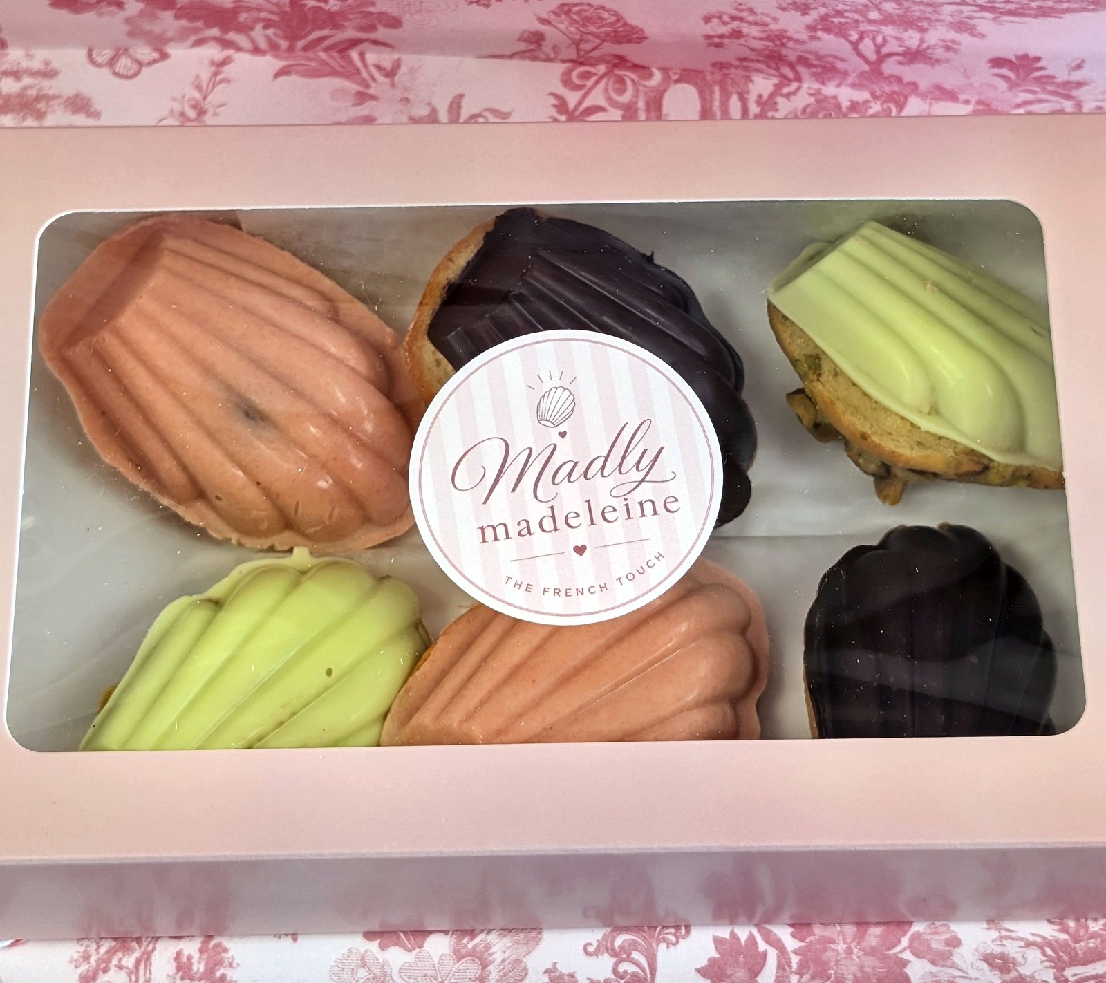 A pink Madly Madeleine box of six madeleines in peach, green and dark chocolate with the logo sticker