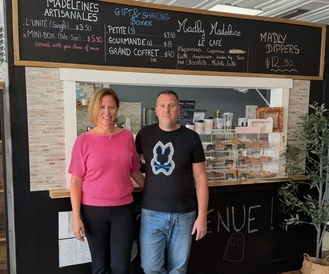 Sabrina and Arnaud standing together at the counter of Madly Madeleine, with the chalkboard menu behind them