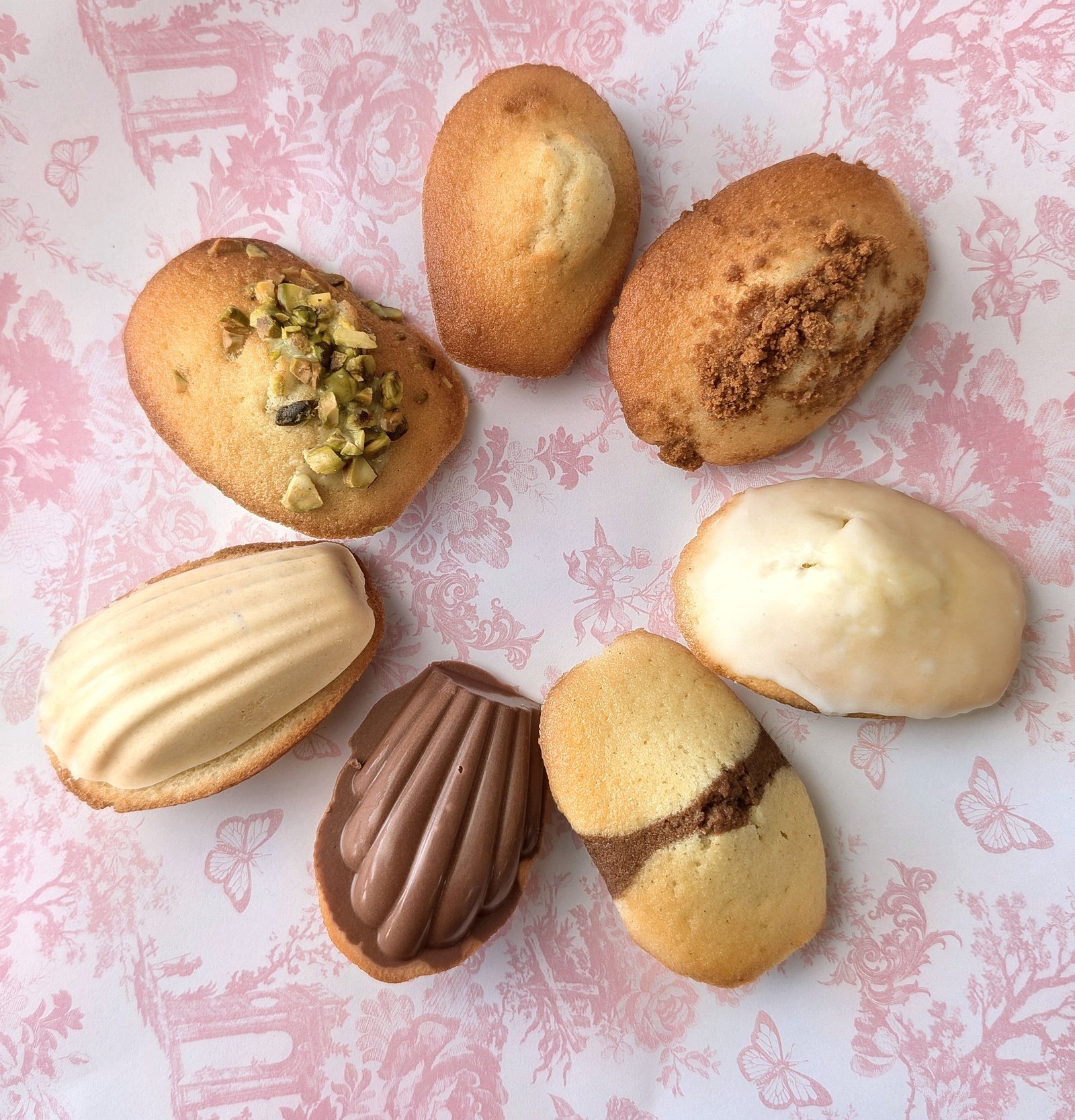 Madeleines in assorted flavors on pink floral paper: pistachio, chocolate-dipped, cookie-crumble and white-glazed