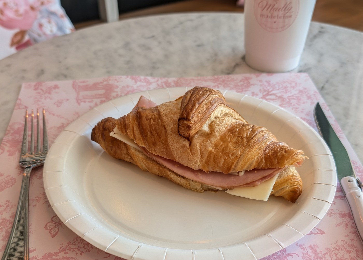 A ham and cheese croissant on a white plate on a pink floral placemat, with a coffee cup on a marble table