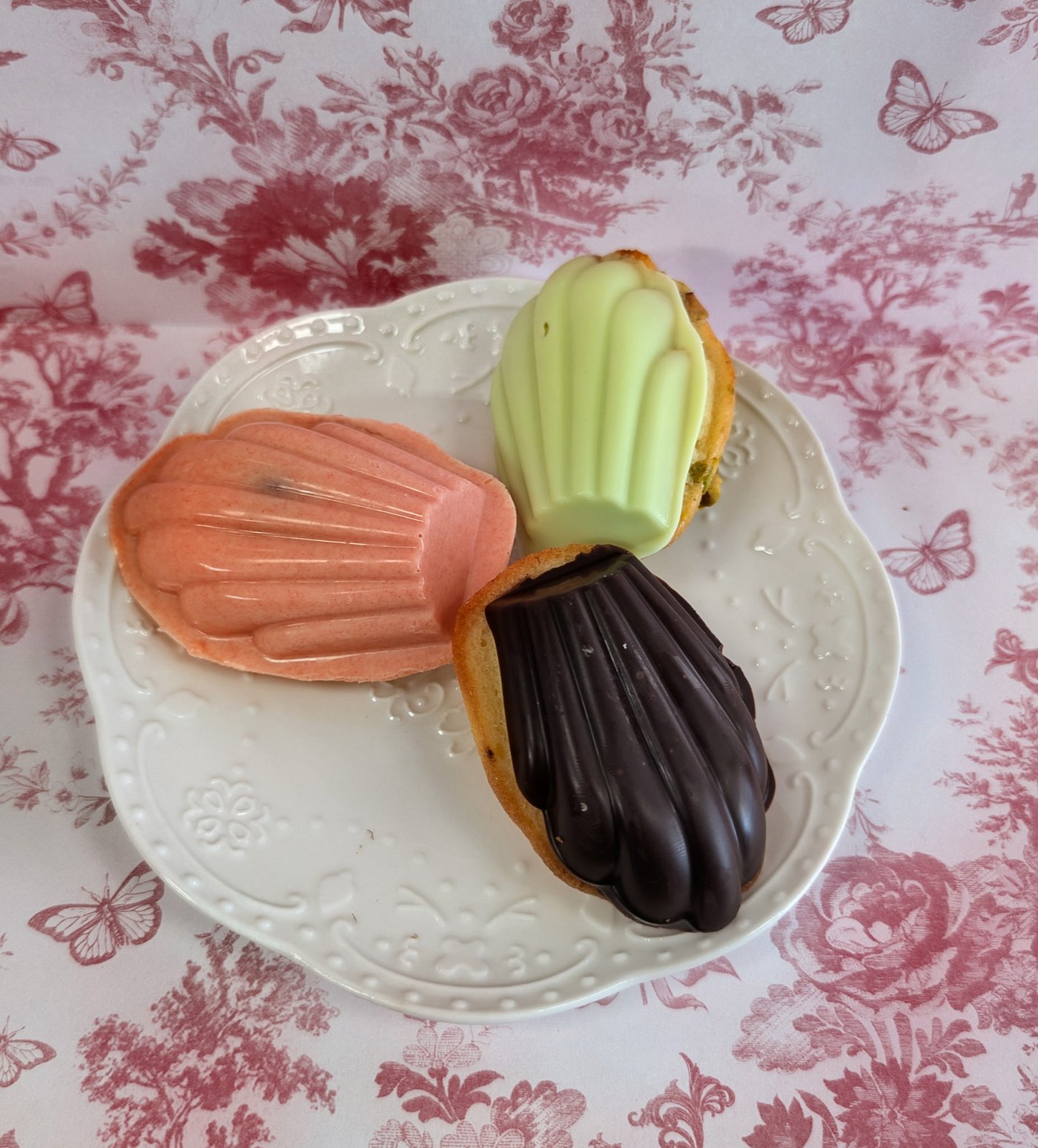 Three chocolate-dipped madeleines in peach, pistachio green and dark chocolate on a white scalloped plate, on pink floral paper