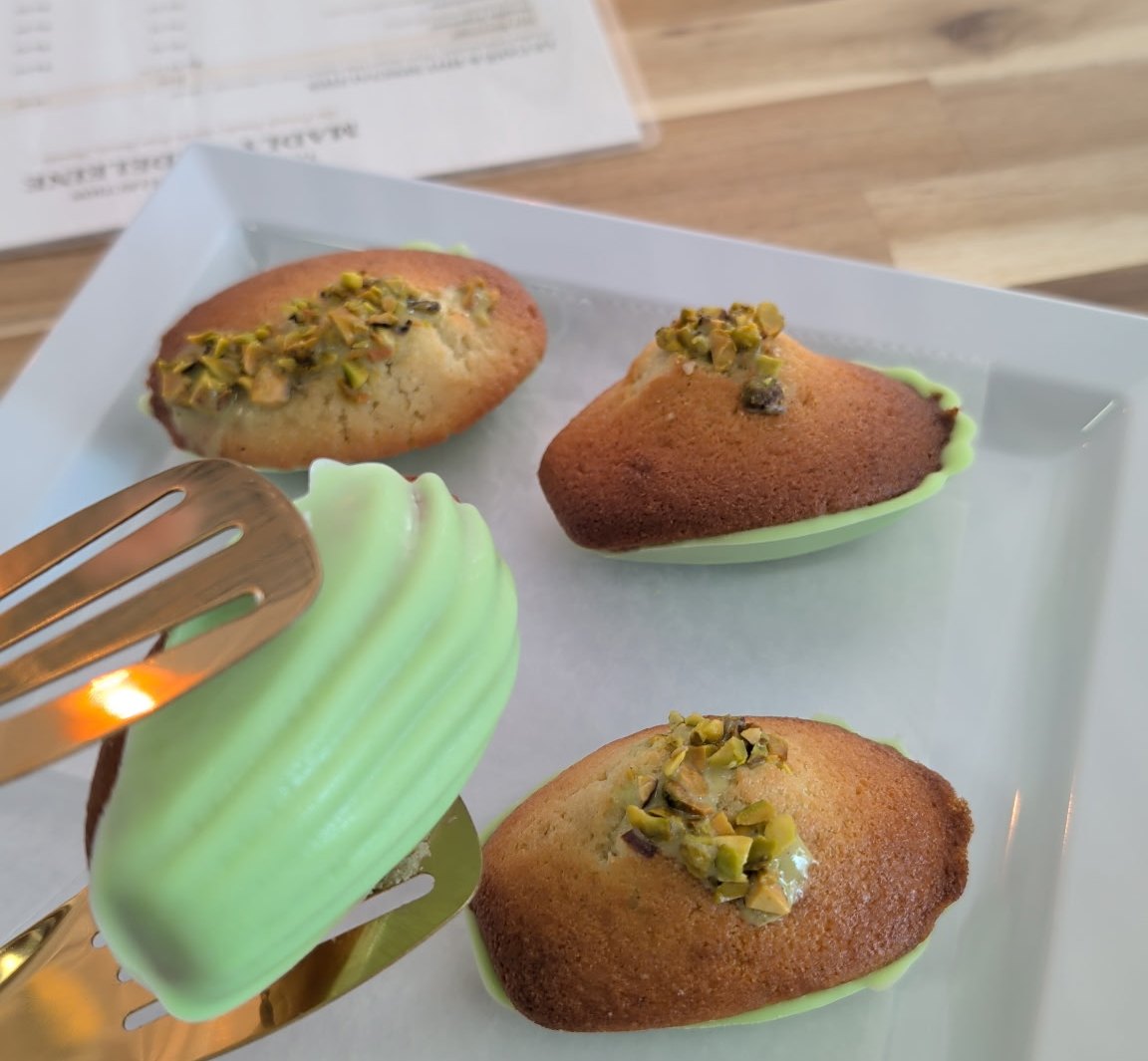 Pistachio madeleines and a green-dipped madeleine on a white tray with gold tongs