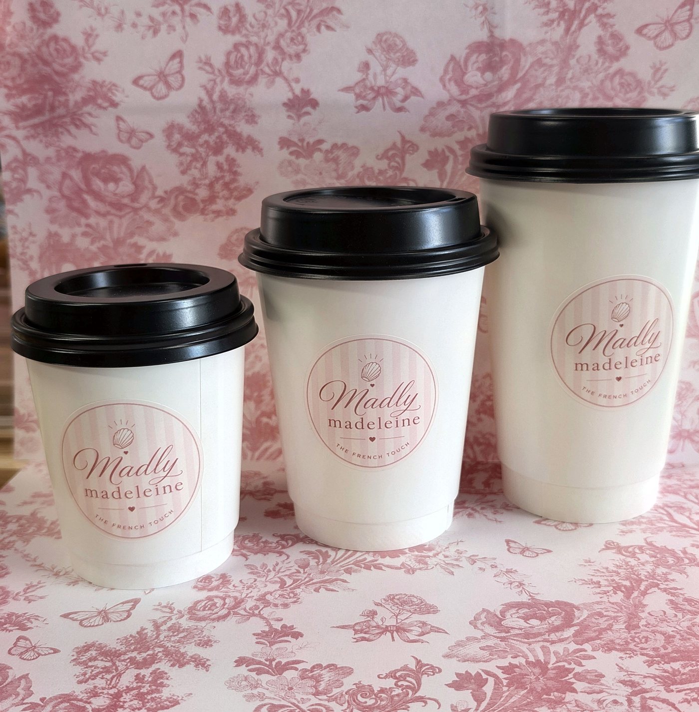 Three Madly Madeleine coffee cups in small, medium and large sizes, each with the logo, on pink floral paper